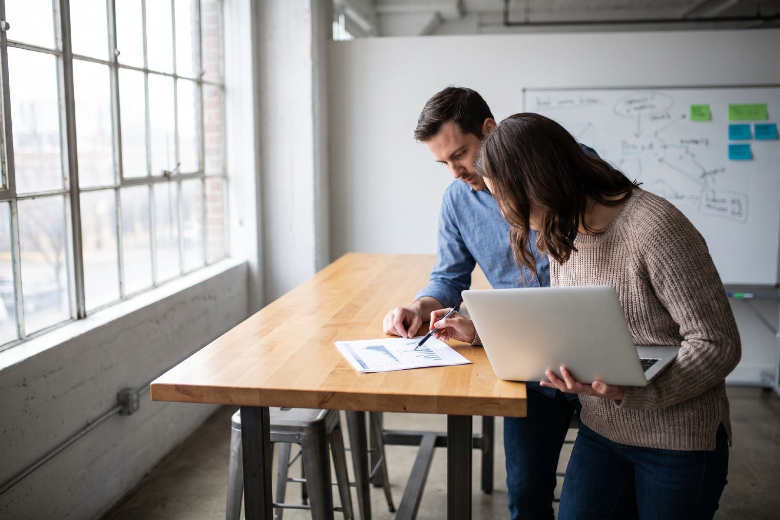 Two people at a high work table reviewing a printed page together, one holding a laptop, a whiteboard behind them.