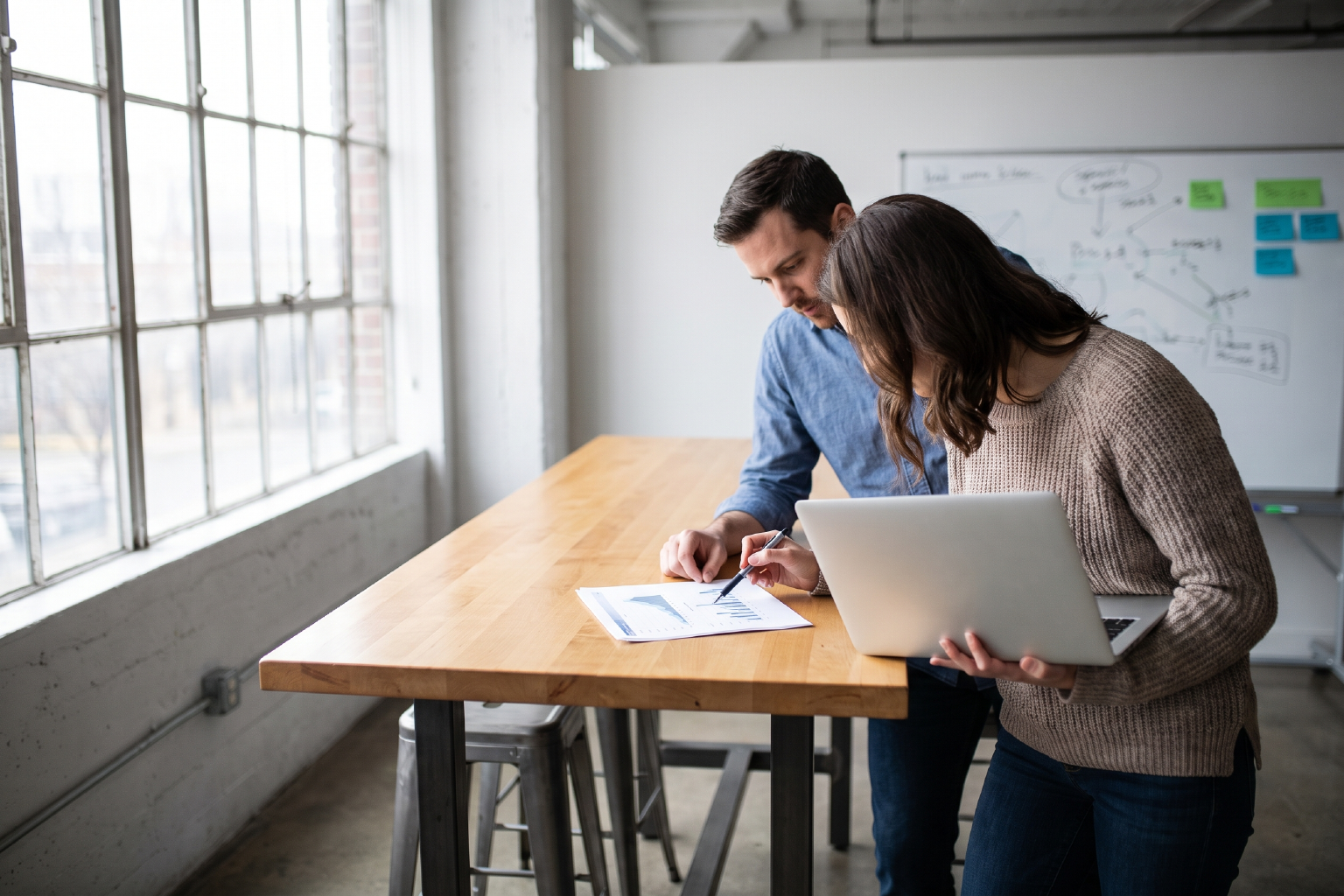 Two people at a high work table reviewing a printed page together, one holding a laptop, a whiteboard behind them.